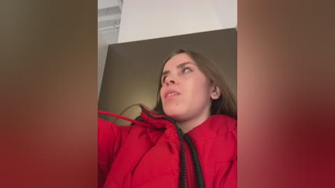 Video of a young Caucasian woman with light skin and long brown hair, wearing a bright red jacket, looking through a rectangular opening.