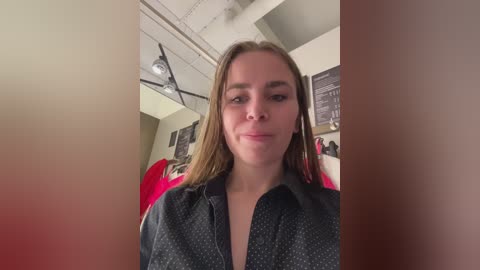 A video of a young woman with straight brown hair, fair skin, and a neutral expression, wearing a black shirt with small polka dots. Background includes a modern, beige-walled room with blackboards, a ceiling light fixture, and people in red clothing.