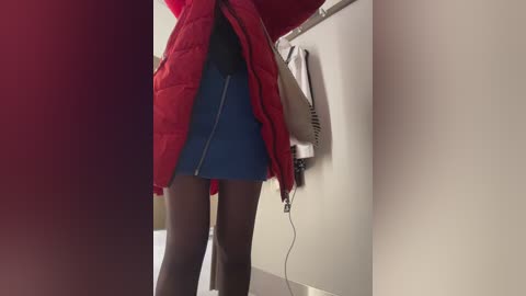 A video of a woman's legs in a red quilted jacket, black tights, and a blue mini skirt, standing next to a beige washing machine with a white towel on top.
