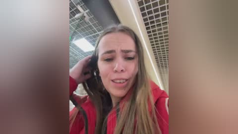 Video of a young woman with long blonde hair, wearing a red jacket, looking concerned while holding her hair. Background features a grid-patterned ceiling and fluorescent lights.