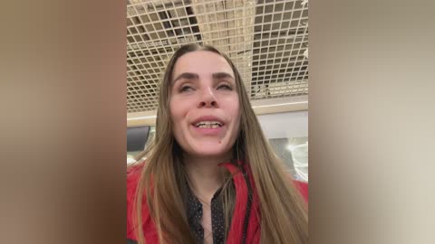 Video of a young Caucasian woman with long blonde hair, smiling broadly, wearing a red jacket. She is indoors with a beige grid-patterned ceiling.