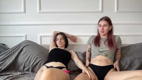 Video of two women in black lingerie on a gray bed, white-paneled wall background, casual and intimate setting.