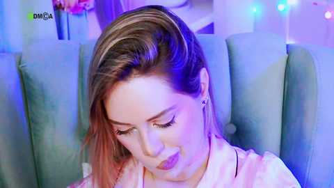 Video of a smiling, light-skinned woman with light brown hair, wearing a pink top, seated on a blue velvet couch. Background includes string lights and a blurred pink flower arrangement.