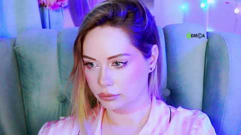 A video of a fair-skinned woman with blonde hair, wearing a pink blouse, sitting in a blue armchair, with soft blue and purple lighting in the background.