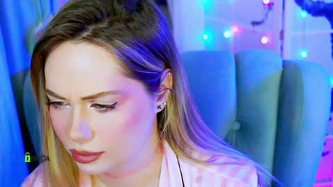 Video of a fair-skinned, blonde-haired woman with a serene expression, wearing a white blouse, against a vibrant blue backdrop with colorful lights.