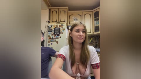A video of a young woman with long blonde hair and fair skin, wearing a low-cut white top with red plaid sleeves, sitting in a cluttered kitchen with a refrigerator full of photos.