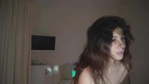 A video of a young woman with long, disheveled black hair and pale skin, partially visible, standing in a dimly lit room with a TV, beige curtains, and a green light.