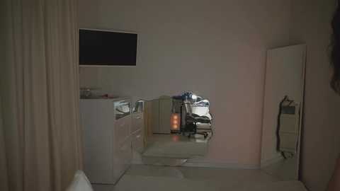 A dimly lit video of a small, cluttered room with beige walls, a white cabinet, a large mirror, a TV, and a partially visible person.