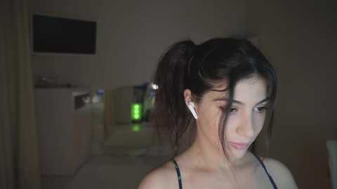 Video of a young woman with fair skin and dark hair tied in a ponytail, wearing a black spaghetti-strap top, listening to white earbuds. Background features dimly lit bedroom with a TV, green glow, and minimalistic decor.