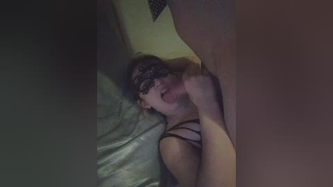 A dimly lit video captures a woman with black lace mask, glasses, and black lingerie, performing oral sex on a man's erect penis, lying on a bed with grey sheets.