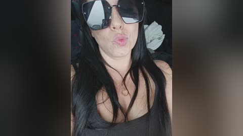 Video of a woman with long black hair, wearing large dark sunglasses and a black tank top, puckering her lips for a kiss.