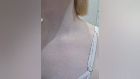 A close-up video of a fair-skinned woman's upper torso, showing her left shoulder, collarbone, and a small mole. She wears a white spaghetti-strap top. The background is blurred, suggesting an indoor setting.