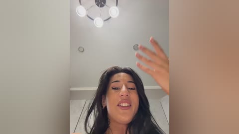 Video of a young woman with long dark hair, light skin, and full lips, captured from below, showing a close-up of her face and raised hand in a modern, white-walled room with a chandelier.