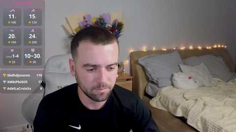 A video of a man with a short beard and a floral crown, sitting on a bed with a white teddy bear, wearing a black shirt. The background features a digital weather display.