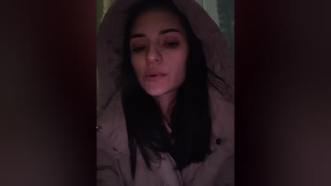 A dimly lit video shows a young woman with long, dark hair, wearing a beige jacket, standing in a dark doorway, her face partially obscured by her arms.