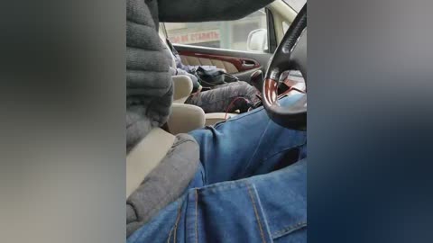 Video of a person sitting in a car, wearing a gray sweater, with blue jeans draped over their lap, showcasing the car's interior with beige seats and a wooden steering wheel.