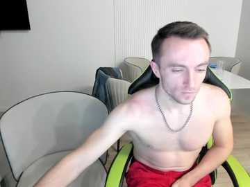 Video of a shirtless, fair-skinned man with short brown hair, wearing red shorts, sitting in a green gaming chair in a modern, minimalist room with white walls and light wood flooring.