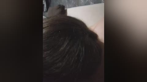 Video of a person's dark brown hair, slightly parted, against a blurred, dark background, with a visible edge of a light-colored object or surface in the upper left corner.