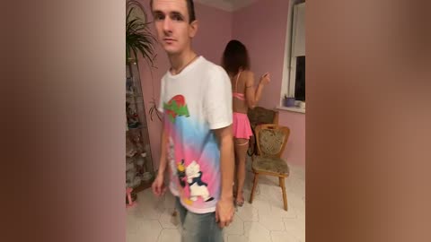 Video of a young man in a colorful graphic t-shirt walking in a pink-painted room with a woman in a pink dress, both looking ahead.