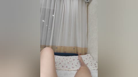 A video showing a person lying on a bed with white floral sheets, legs up, taken from below. The background features sheer white curtains with wooden trim, and a white wall with a mounted air conditioner.