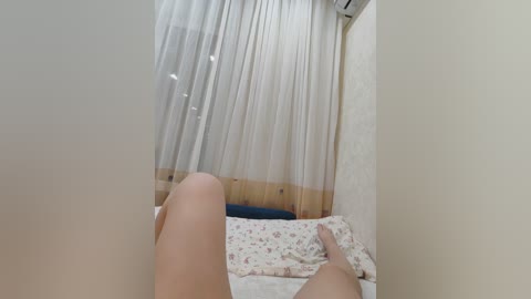 Video of a person lying on a bed, legs extended, in a dimly lit room with sheer curtains covering a window.