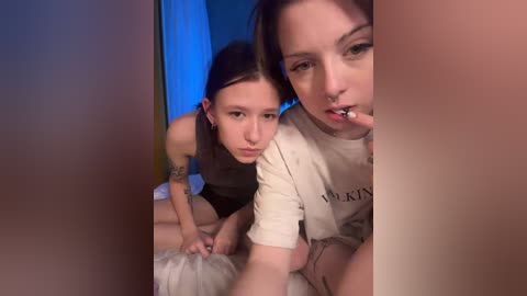 Video of two young women with pale skin, dark hair, and tattoos, wearing casual clothes, sitting on a bed with blue curtains in the background.