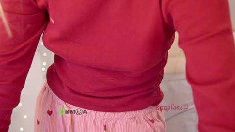 Video of a woman's torso in a red sweater, pink striped pajama bottoms, with a blurred background featuring soft lighting and a bed.