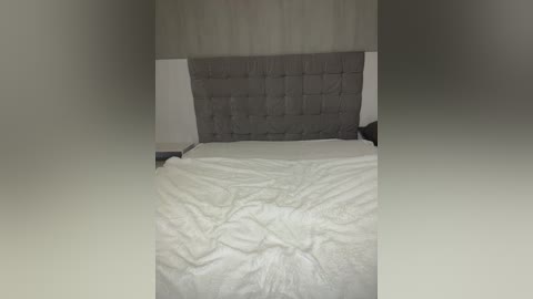 A video of a minimalist bedroom featuring a neatly made bed with white linens and a gray tufted headboard against a plain, beige wall.