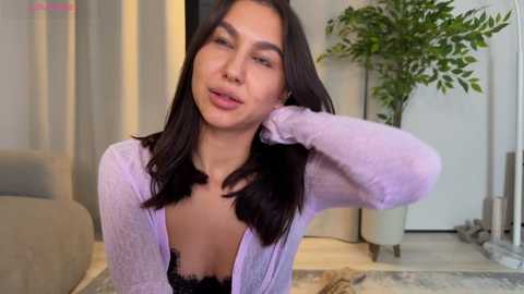 Video of a young woman with medium-length black hair, wearing a lavender cardigan and black lace bra, posing in a modern living room with a potted plant and beige furniture.