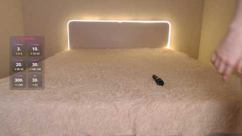 A video of a plush, cream-colored bed with a black vibrator on the bedspread. A woman's hand is reaching toward the vibrator. The room is dimly lit with a warm, glowing light.