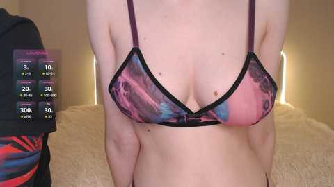A video of a woman in a sheer, colorful, patterned bra with black straps, standing indoors with a blurred background. A digital display on the left shows health stats.