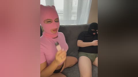 Video: A woman in a pink mask and tight top, holding a fork, sits next to a man in a black mask, both on a gray sofa.