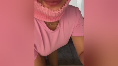 A close-up video of a woman's face and upper chest in a pink sweater and necklace, partially obscured by pink fabric, with a gray skirt in the background.