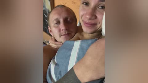 A video shows a smiling, middle-aged, fair-skinned woman with short blonde hair, wearing a blue-striped top, hugging a bald, fair-skinned man with a neutral expression. The background features wooden walls and a framed picture.