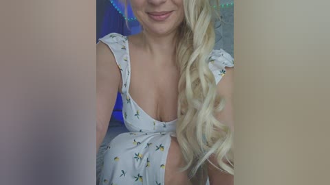 Video of a smiling, blonde woman with long, curly hair, wearing a white sundress with yellow and green floral patterns. She is indoors, with a blurred background of blue lights and a light-colored wall.