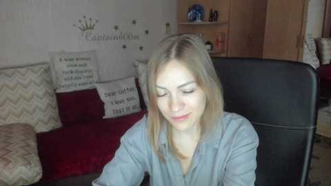 A video of a blonde woman with fair skin, wearing a blue shirt, smiling, seated on a black leather chair in a cozy living room with wooden furniture and decorative pillows.