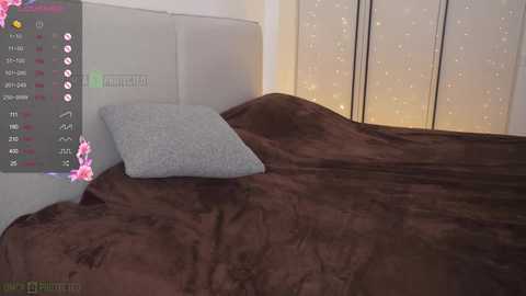 A video of a cozy bedroom featuring a bed with a plush, dark brown blanket, a light gray pillow, and a white padded headboard. A digital overlay displays gaming stats in the corner.