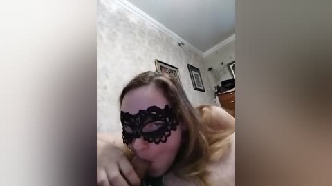 Video of a light-skinned woman with shoulder-length brown hair, wearing a black lace mask, performing oral sex on a man in a dimly lit room with floral wallpaper and framed photos on the wall.