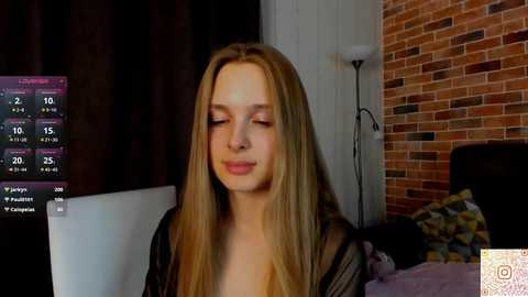 A video of a young Caucasian woman with long blonde hair, wearing a sheer black robe, sitting indoors with a brick wall and a modern lamp in the background.