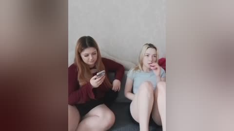 A video featuring two young women, one brunette in a maroon sweater and shorts, and another blonde in a light blue shirt, sitting on a sofa with a blurred background.