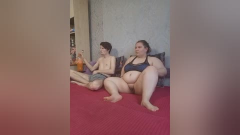 Video of two overweight people, a man and a woman, sitting on a bed with red sheets. They are wearing casual clothing and drinking from a bottle. The room has a beige wall and a bookshelf in the background.
