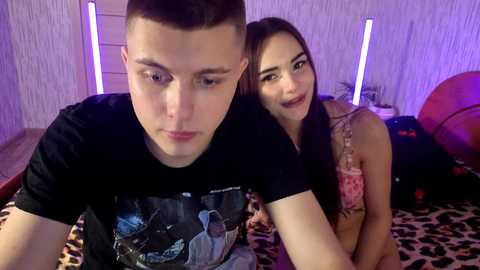 Video of a young Caucasian man and woman, both with light skin, sitting on a leopard-print bed in a dimly lit room with purple lighting. The man wears a black T-shirt, and the woman is topless, smiling.