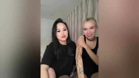 Video of two women with long black hair, one wearing a black shirt, the other a black tank top, in a bedroom with beige curtains and a bed.