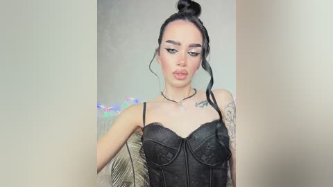 Video of a pale-skinned woman with dark hair in a topknot, wearing a black lace corset and a tattoo on her shoulder, standing against a plain beige wall.