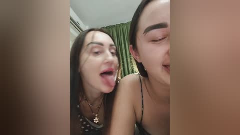 Video of two young women with fair skin, one sticking out her tongue, wearing a black spaghetti strap top, and the other with her eyes closed, also wearing a black top. Green curtains in the background.
