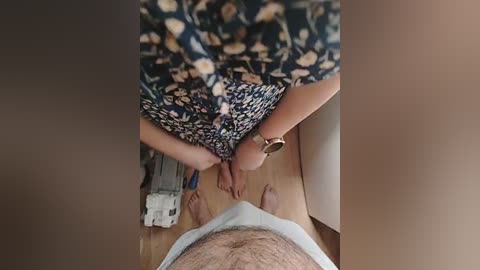 Video: Woman in floral dress kneeling beside a man's bare legs, wooden floor, white appliance in background.