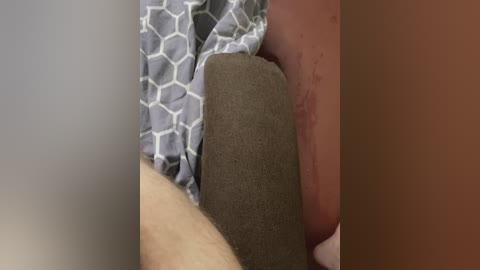 Video of a close-up scene with a hand holding a grey, textured object, possibly a pillow, against a person's bare, lightly tanned thigh. The background shows a grey, geometric-patterned blanket.