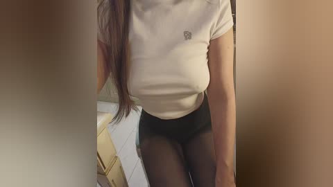 A video of a woman in a tight white t-shirt and black pantyhose, taken from a slightly blurred overhead angle. Her long brown hair falls over her shoulder. The background features a tiled bathroom.