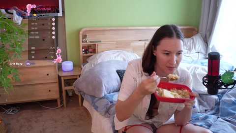 Video of a young woman with long brown hair, wearing a white robe, eating from a red bowl, sitting on a bed with a wooden headboard, in a cozy, green-painted room.