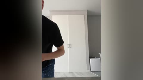 Video of a person in a black t-shirt, standing in front of a minimalist, white wardrobe and a white washing machine in a modern, light gray room.